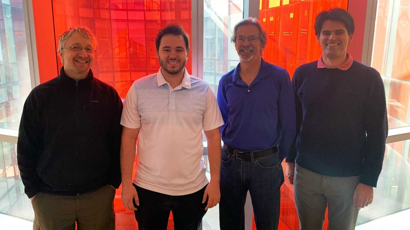 Four men stand in a row in front of alternating white and red windows.